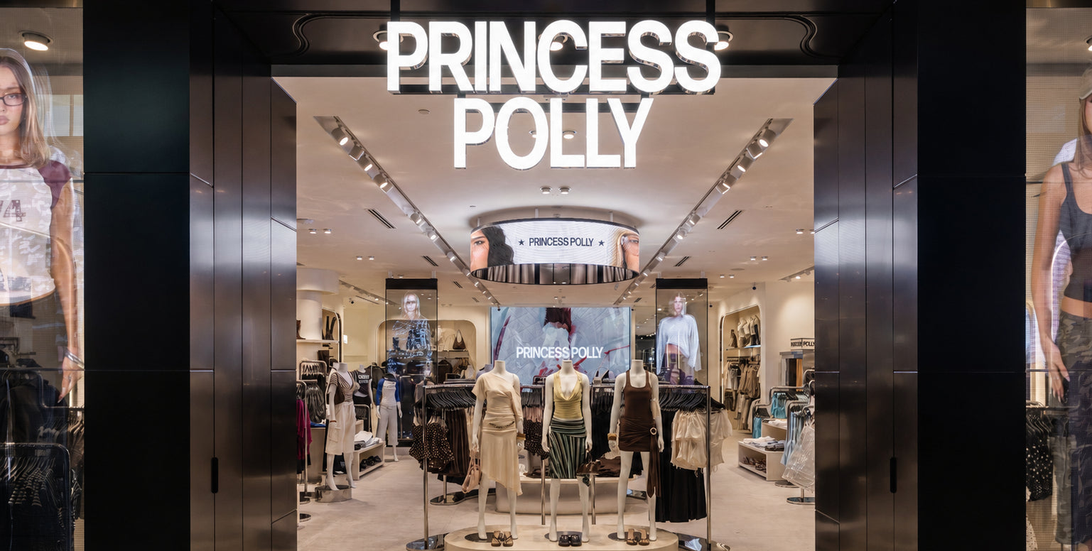 Princess Polly clothing store entrance with illuminated white brand signage above a wide opening, showing mannequins and racks of apparel inside.